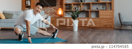 Happy mature sportsman making exercises for legs, stretching on fitness mat in living room interior and smiling at camera. Home training, workout, fitness and weight loss, copy space 114704585