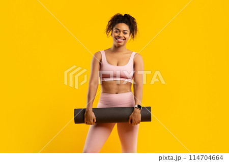 Smiling young african american woman in sportswear holding a yoga mat, set against a vibrant yellow background 114704664