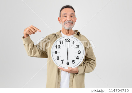 A cheerful older man with a beard is holding a large wall clock and pointing at it, standing against a plain background A cheerful older man with a beard is holding a large wall clock and pointing at it, standing against a plain background 114704730