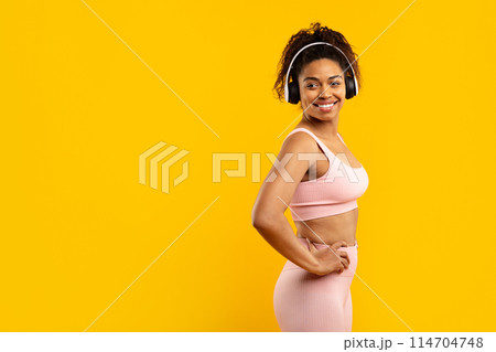 Fit black lady confidently poses with hands on hips, in workout clothing and headphones on yellow, copy space 114704748