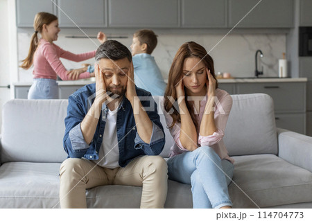 A stressed couple sits on a couch, looking exhausted, while their two children fight in the background of a modern home 114704773