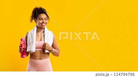 Radiant african american woman in fitness attire holding a pink water bottle, smiling with a towel around her neck on a yellow background Radiant african american woman in fitness attire holding a pink water bottle, smiling with a towel around her neck on a yellow background 114704798