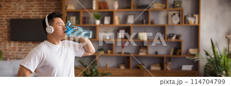 Sporty mature korean man with headphones drinking water after workout at home, staying hydrated during domestic sports training. Healthy lifestyle, wellness concept, copy space 114704799