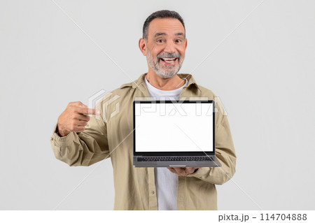 Cheerful mature man presenting a laptop with a blank screen, dressed casually, standing against a grey background 114704888
