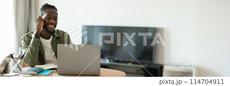 Remote career. Happy african american man talking on cellphone and using laptop, sitting at workplace at home. Freelancer guy having phone call and working on pc computer, copy space, banner 114704911
