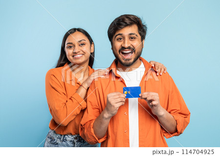 Cheerful Indian couple in orange shirts holding a credit card, suggesting shopping or payment on blue 114704925