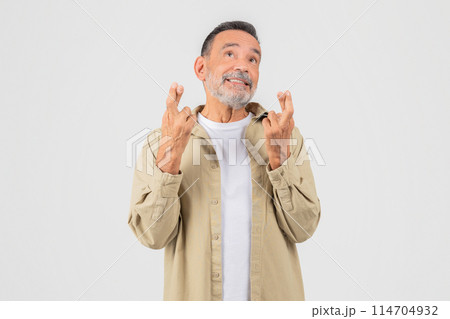 A hopeful old man with a beard is looking up, fingers crossed, wearing a beige shirt and white t-shirt on an isolated background Includes keywords like senior, elderly, old, man, isolated 114704932