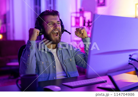 A man with a beard is laughing while gaming, illuminated by the glow of a computer screen in a dark room A man with a beard is laughing while gaming, illuminated by the glow of a computer screen in a dark room 114704986