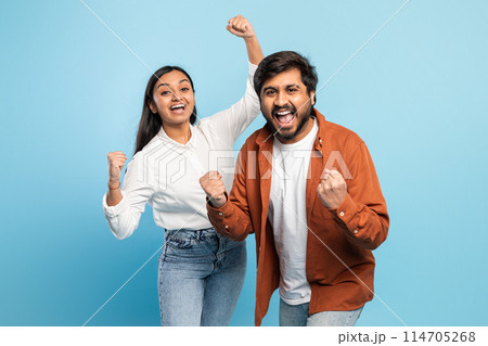 A joyful indian couple celebrates with fists raised in a victory or success gesture against a blue backdrop A joyful indian couple celebrates with fists raised in a victory or success gesture against a blue backdrop 114705268