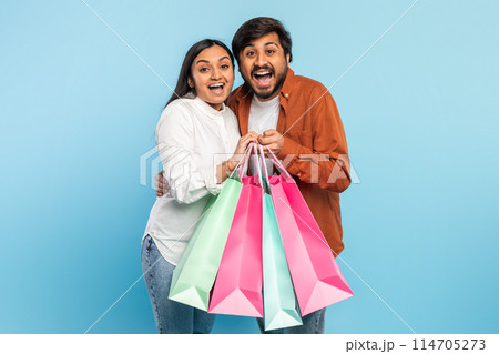 An ecstatic indian couple holds shopping bags up high, expressing excitement with wide smiles on a blue backdrop An ecstatic indian couple holds shopping bags up high, expressing excitement with wide smiles on a blue backdrop 114705273