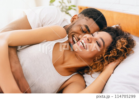 Happy millennial black couple flirting in morning on comfortable bed, tickling and laughing Happy millennial black couple flirting in morning on comfortable bed, tickling and laughing 114705299