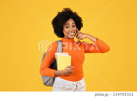 Cheerful young woman with curly hair holding notebooks, making a call me gesture, clad in an orange turtleneck and white pants, with a grey backpack on a sunny yellow background 114705331