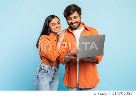 Indian couple in orange attire smiling while collaborating and looking at laptop screen, modern work concept 114705356