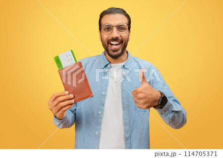 Joyful indian man shows approval with passport and boarding passes on yellow, signaling positive travel experience 114705371