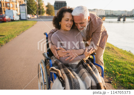 Senior woman in wheelchair walking with caregiver old man on road in park. Elderly family couple man supporting embracing woman in chair for people with disability outdoor. Rehabilitation 114705373
