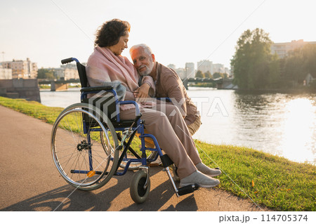 Senior woman in wheelchair walking with caregiver old man on road. Elderly family couple man supporting taking care of paralyzed woman in chair for people with disability outdoor. Rehabilitation 114705374