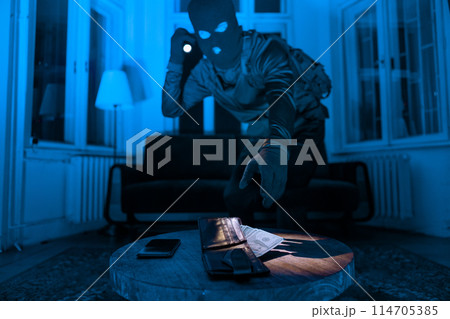 A masked burglar in a dark room uses a flashlight to find and steal a wallet and phone on a wooden table 114705385