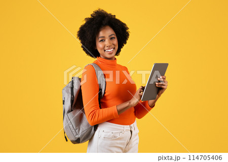 Confident smiling African American woman student with curly hair smiling and holding a digital tablet, with a backpack slung over her shoulder, against a yellow background Confident smiling African American woman student with curly hair smiling and holding a digital tablet, with a backpack slung over her shoulder, against a yellow background 114705406