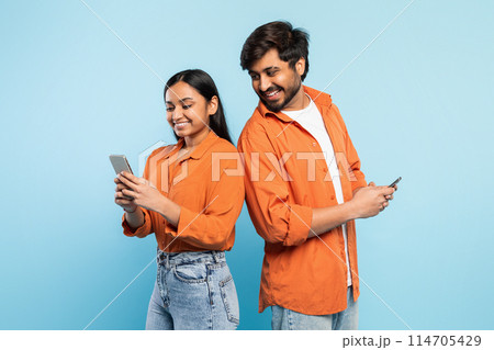 Casual young indian couple using their phones, engrossed in personal digital worlds with blue backdrop Casual young indian couple using their phones, engrossed in personal digital worlds with blue backdrop 114705429