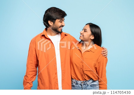 An indian man and woman stand close together in casual orange shirts, making a heartwarming display against a solid blue backdrop, faces blurred An indian man and woman stand close together in casual orange shirts, making a heartwarming display against a solid blue backdrop, faces blurred 114705430