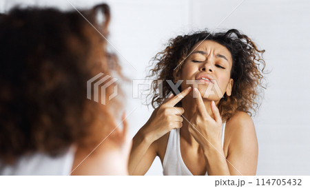 Afro girl frowning squeezing pimple on her face in mirror. Do not squeez pimples concept, copy space Afro girl frowning squeezing pimple on her face in mirror. Do not squeez pimples concept, copy space 114705432
