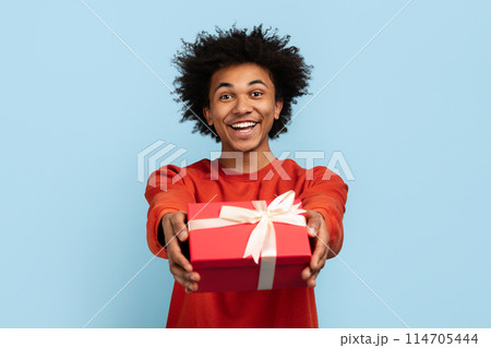 Cheerful african american man holds out a red gift box with a white ribbon, looking friendly 114705444