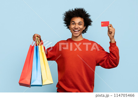 Exuberant african american man holding colorful shopping bags and a credit card on blue background 114705466