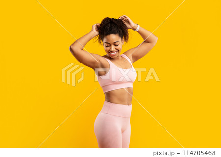 A vibrant young african american woman in pink workout attire happily prepares her hair for a fitness session against a yellow wall A vibrant young african american woman in pink workout attire happily prepares her hair for a fitness session against a yellow wall 114705468