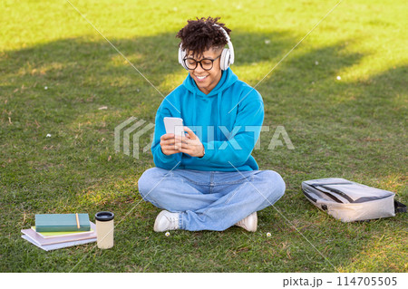 A young brazilian guy student with headphones sits outdoors focusing on his smartphone, surrounded by books and a coffee cup A young brazilian guy student with headphones sits outdoors focusing on his smartphone, surrounded by books and a coffee cup 114705505