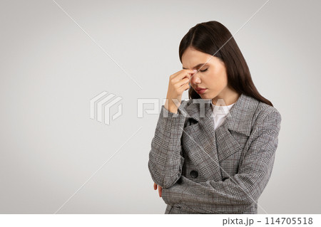 Woman in checkered blazer showing signs of stress with headache, pinching the bridge of her nose, against minimalist gray backdrop, free space 114705518