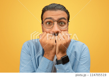 Biting nails, a worried indian man stands looking anxious against a yellow background, signifying tension Biting nails, a worried indian man stands looking anxious against a yellow background, signifying tension 114705519