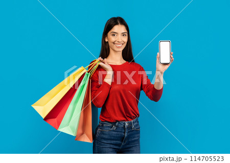 Woman holding multiple shopping bags and showing blank cell phone, happy female recommending new app for purchases, standing on blue studio background, mockup 114705523