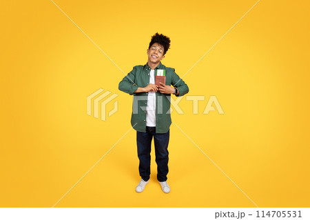 A cheerful young african american guy with curly hair, holding a passport with a boarding pass against a yellow background, full length 114705531