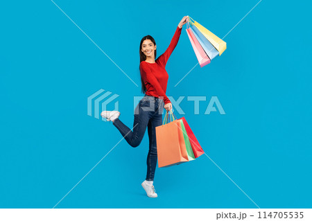 Excited young woman joyfully jumping in the air while holding shopping bags, showcasing her excitement for her purchases, posing on blue background 114705535
