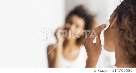 Young afro-american woman cleaning face with cotton pad in front of mirror in bathroom. Daily face cleaning concept 114705580