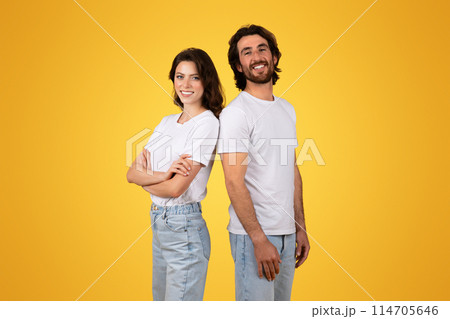 Side-by-side, a confident european millennial woman and a beaming man stand back-to-back in plain white t-shirts and light blue jeans against a sunny yellow backdrop, studio 114705646