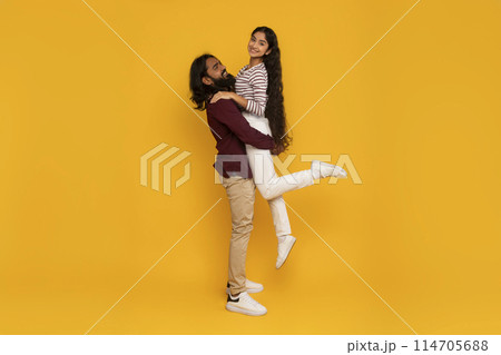 A happy man with a beard is smiling while carrying a woman in a joyful pose against a vivid yellow background A happy man with a beard is smiling while carrying a woman in a joyful pose against a vivid yellow background 114705688