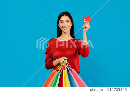 Young happy woman holding credit card in one hand and bright shopper bags in the other, happy female ready for shopping spree, standing on blue studio background, copy space Young happy woman holding credit card in one hand and bright shopper bags in the other, happy female ready for shopping spree, standing on blue studio background, copy space 114705849