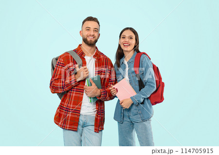 Two happy students man and woman standing with backpacks and books against blue backdrop, smiling to camera, representing concept of modern university life and study. Studio shot 114705915