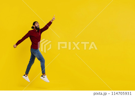 Excited handsome bearded young indian man hipster jumping in the air isolated on yellow studio background, pointing at copy space, showing great online offer or amazing deal, full length 114705931