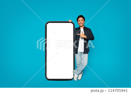 Confident millennial japanese man in a casual dark jacket and jeans standing next to a giant smartphone with a blank display, pointing at it with a friendly smile against a striking blue background Confident millennial japanese man in a casual dark jacket and jeans standing next to a giant smartphone with a blank display, pointing at it with a friendly smile against a striking blue background 114705941