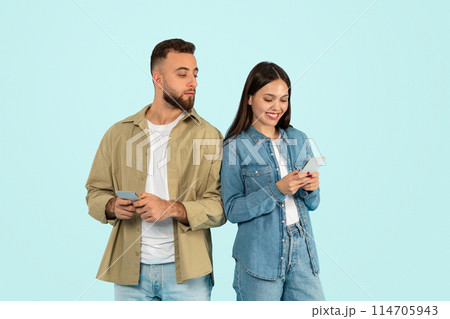 Jealousy issue. Distrustful nosy guy sneakily peeks at his girlfriend's phone, reading her messages behind her back, suspecting cheating standing together with gadgets on blue backdrop Jealousy issue. Distrustful nosy guy sneakily peeks at his girlfriend's phone, reading her messages behind her back, suspecting cheating standing together with gadgets on blue backdrop 114705943
