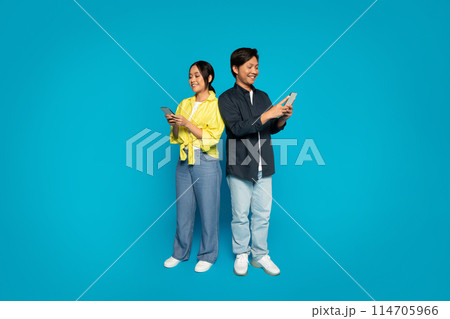 A japanese millennial man and woman stand back to back, each engrossed in their own smartphones, dressed in casual clothing with pleasant expressions against a bright blue background A japanese millennial man and woman stand back to back, each engrossed in their own smartphones, dressed in casual clothing with pleasant expressions against a bright blue background 114705966