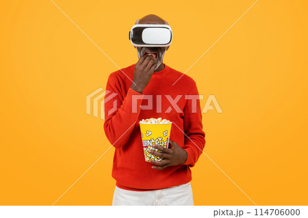 Engrossed senior black man in a red sweater enjoying virtual reality with a headset, snacking on popcorn, and displaying a look of surprise on a yellow background, studio Engrossed senior black man in a red sweater enjoying virtual reality with a headset, snacking on popcorn, and displaying a look of surprise on a yellow background, studio 114706000