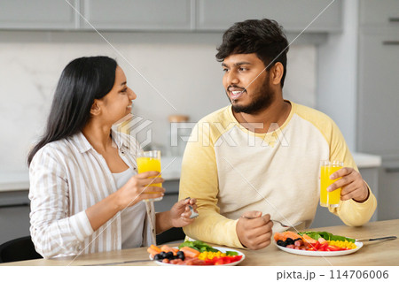 Positive young indian spouses enjoying breakfast together at home. Eastern loving couple have meal, drinking fresh orange juice, chatting and smiling, kitchen interior 114706006