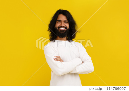 Confident handsome bearded long-haired millennial indian guy wearing white long sleeve posing with arms crossed on chest, smiling at camera, isolated on yellow studio background 114706007