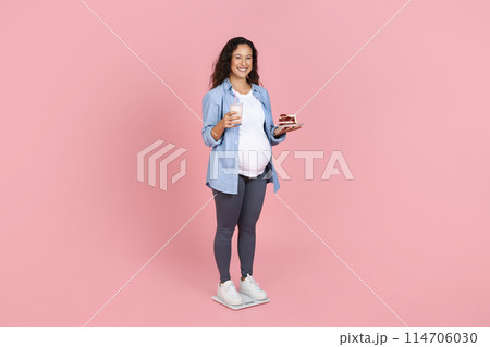 Happy young expecting lady holding glass of milk and cake, have snack, feeling hungry, isolated on pink studio background, copy space, full length. Unhealthy eating during pregnancy 114706030