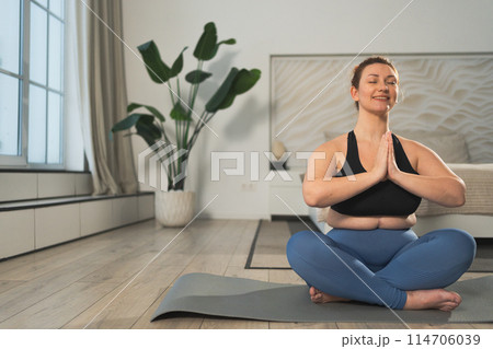 Yoga mindfulness meditation. Woman practicing yoga in living room at home. Plus size female sitting in lotus pose meditating smiling relaxing indoor. Girl doing breathing practice. Yoga at home 114706039
