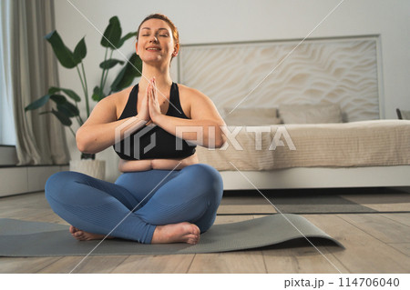 Yoga mindfulness meditation. Woman practicing yoga in living room at home. Plus size female sitting in lotus pose meditating smiling relaxing indoor. Girl doing breathing practice. Yoga at home 114706040