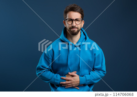 Stomach Pain. Sick young man suffering from acute abdominal ache, upset millennial guy touching belly and frowning, feeling unwell while standing against blue studio background, copy space 114706068
